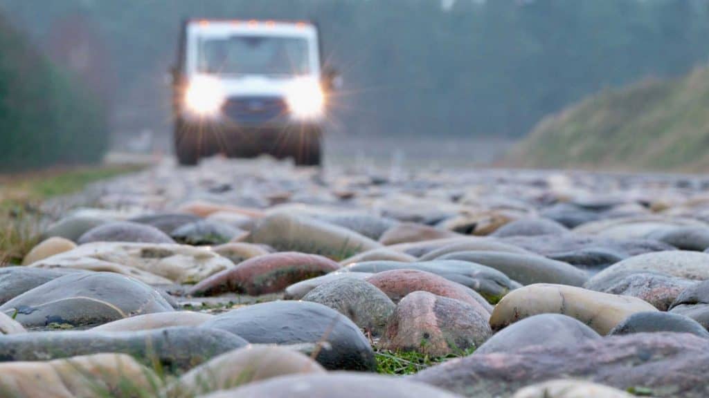Imagem mostra, em destaque, pista bastante irregular. Ao fundo, um carro em movimento.