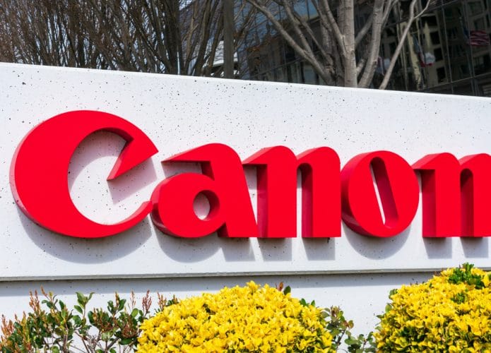 Imagem mostra o logotipo da empresa Canon, em um totem rodeado por plantas