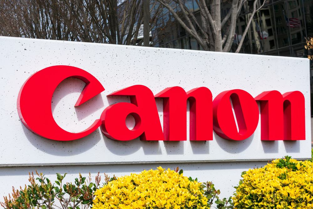 Imagem mostra o logotipo da empresa Canon, em um totem rodeado por plantas