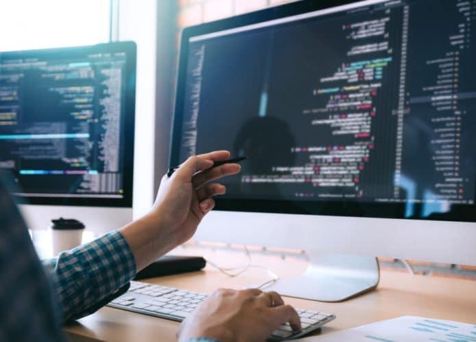 Imagem mostra um computador em cima de uma mesa; no monitor aparecem códigos de programação e, à frente, há uma pessoa apontando para a tela com uma caneta