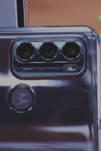 Imagem mostra a parte traseira do Moto G60, evidenciando suas três câmeras, eum boneco de corda do robô do Android ao fundo