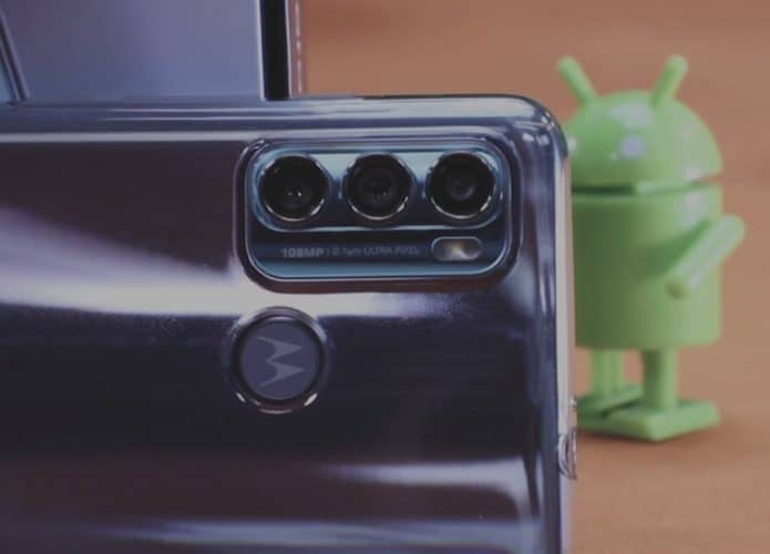 Imagem mostra a parte traseira do Moto G60, evidenciando suas três câmeras, eum boneco de corda do robô do Android ao fundo