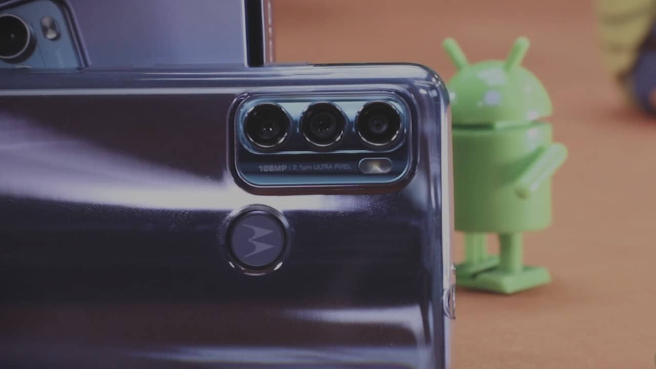 Imagem mostra a parte traseira do Moto G60, evidenciando suas três câmeras, eum boneco de corda do robô do Android ao fundo