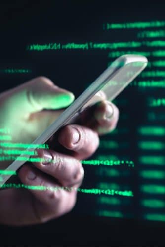 Imagem mostra uma mão segurando um smartphone e, ao redor dela, uma série de códigos e dados em fonte verde, simbolizando o acesso a informações privadas e golpes cibernéticos