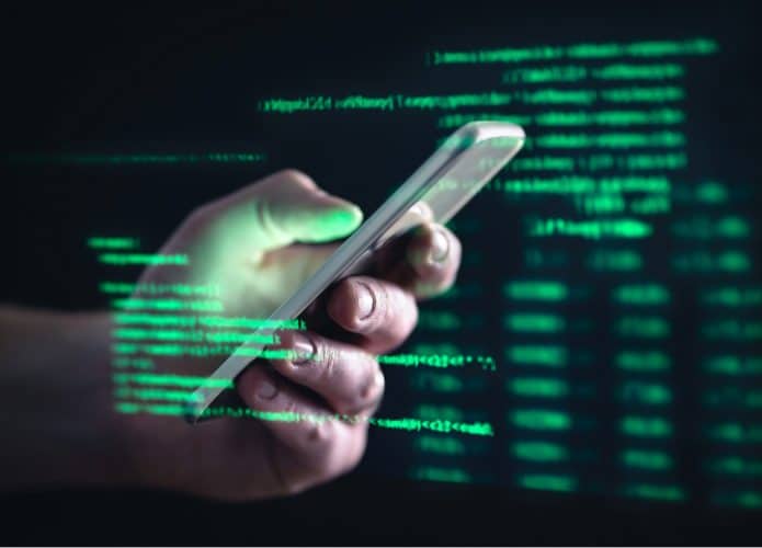 Imagem mostra uma mão segurando um smartphone e, ao redor dela, uma série de códigos e dados em fonte verde, simbolizando o acesso a informações privadas e golpes cibernéticos