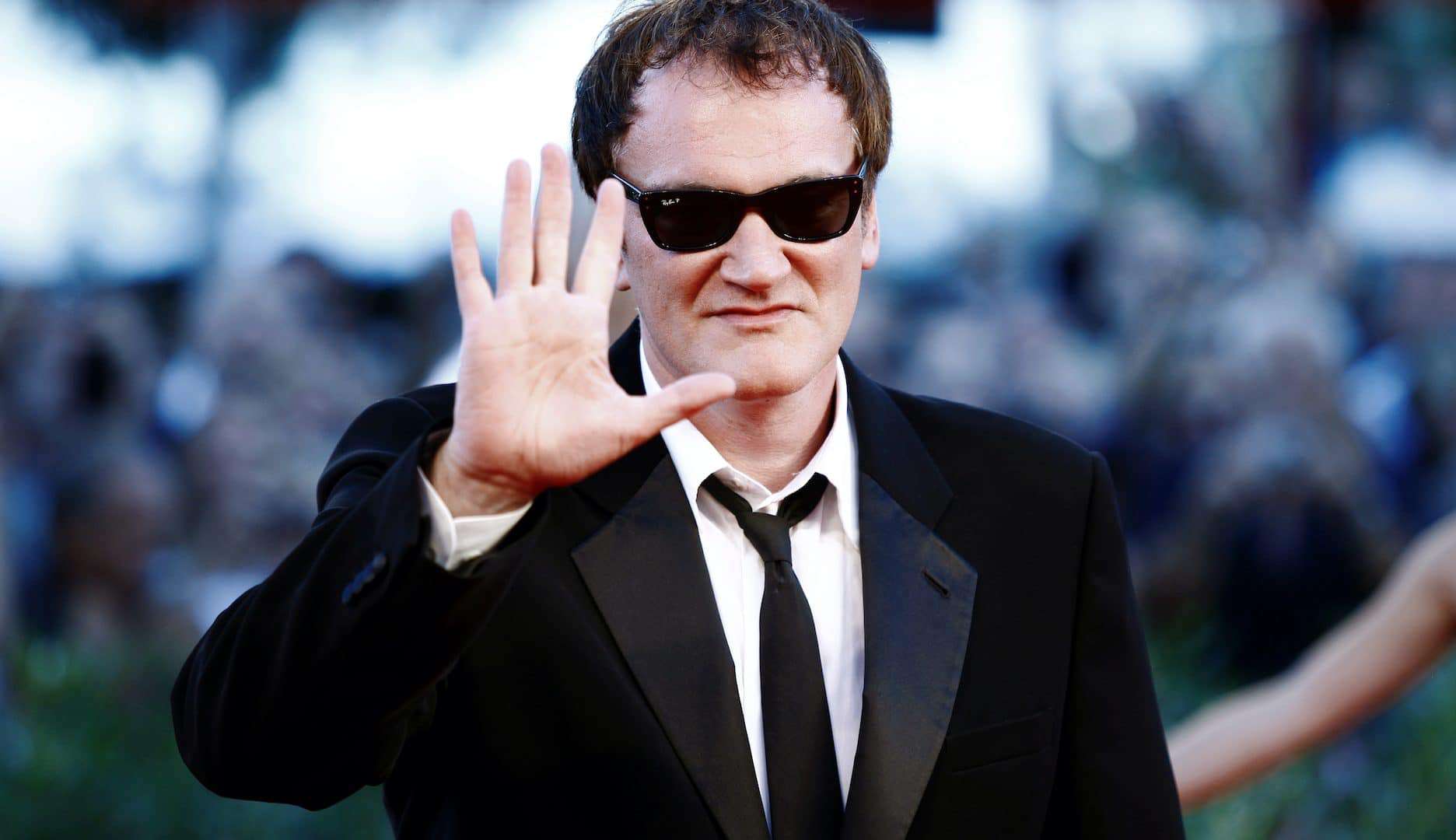 Quentin Tarantino de fraque e óculos escuro exibindo a palma da mão.