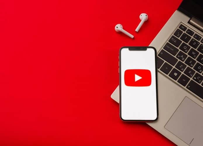 Imagem mostra logo do youtube na tela de um smartphone que, por dua vez, encontra-se em cima de um teclado de notebook. O fundo da imagem é vermelho