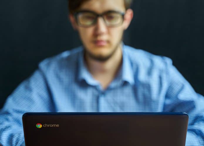 Imagem mostra um homem usando um Chromebook. O homem está desfocado ao fundo da foto, com a tampa do computador e o logotipo do Chrome OS em evidência na frente