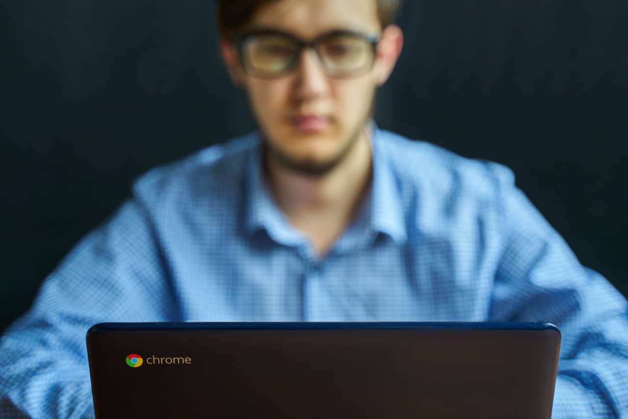 Imagem mostra um homem usando um Chromebook. O homem está desfocado ao fundo da foto, com a tampa do computador e o logotipo do Chrome OS em evidência na frente