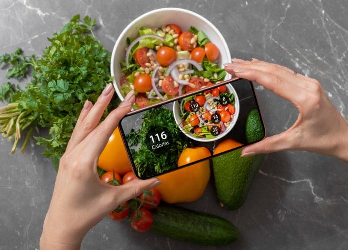 Imagem mostra um prato de salada sob um fundo cinza de pedra. À frente, duas mãos femininas seguram um smartphone, que está tirando foto da comida