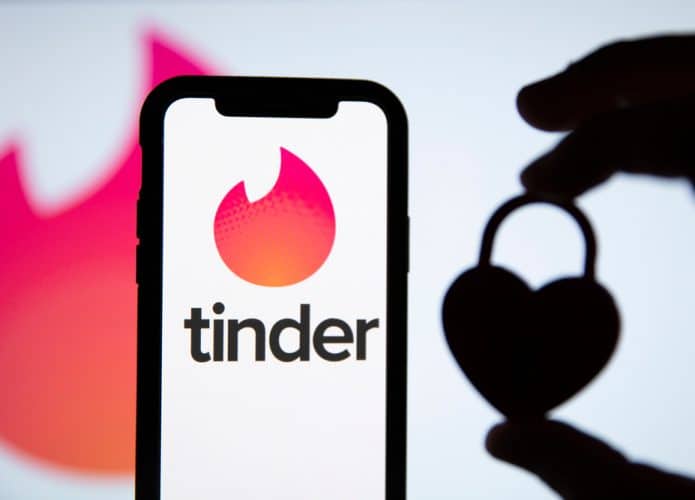 Ao fundo é possível ver a logomarca do Tinder, à frente aparece uma mão segurando um cadeado em formato de coração à direita; à esquerda está um smartphone com o logo do Tinder na tela
