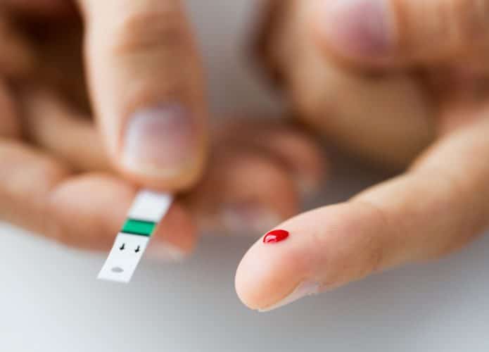 Imagem mostra um dedo perfurado, com uma gota de sangue à mostra. Na outra mão, um teste de diabetes