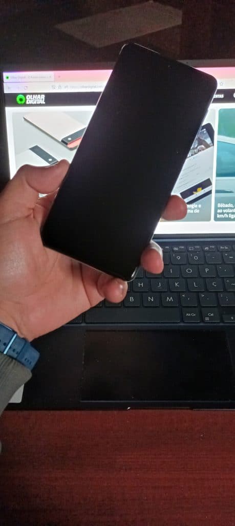 Imagem mostra a tela da frente do Galaxy M62, sendo segurado por uma mão e um notebook exibindo a página inicial do Olhar Digital ao fundo. Samsung lançou o Galaxy M32 e o Galaxy M62 no varejo online brasileiro em 2 de agosto