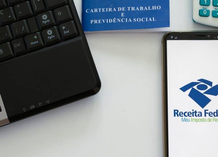 Computador, carteira de trabalho e celular mostrando tela da Restituição do Imposto de Renda