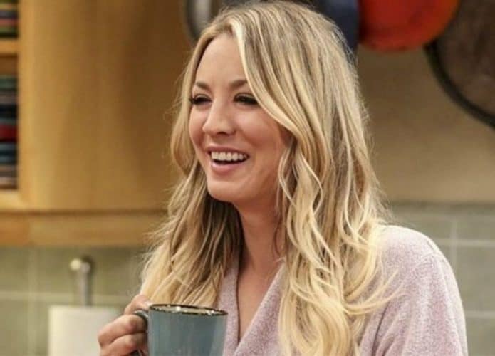 Kaley Cuoco, caracterizada como Penny, segura uma xícara de café e sorri.