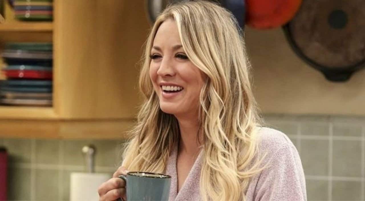 Kaley Cuoco, caracterizada como Penny, segura uma xícara de café e sorri.