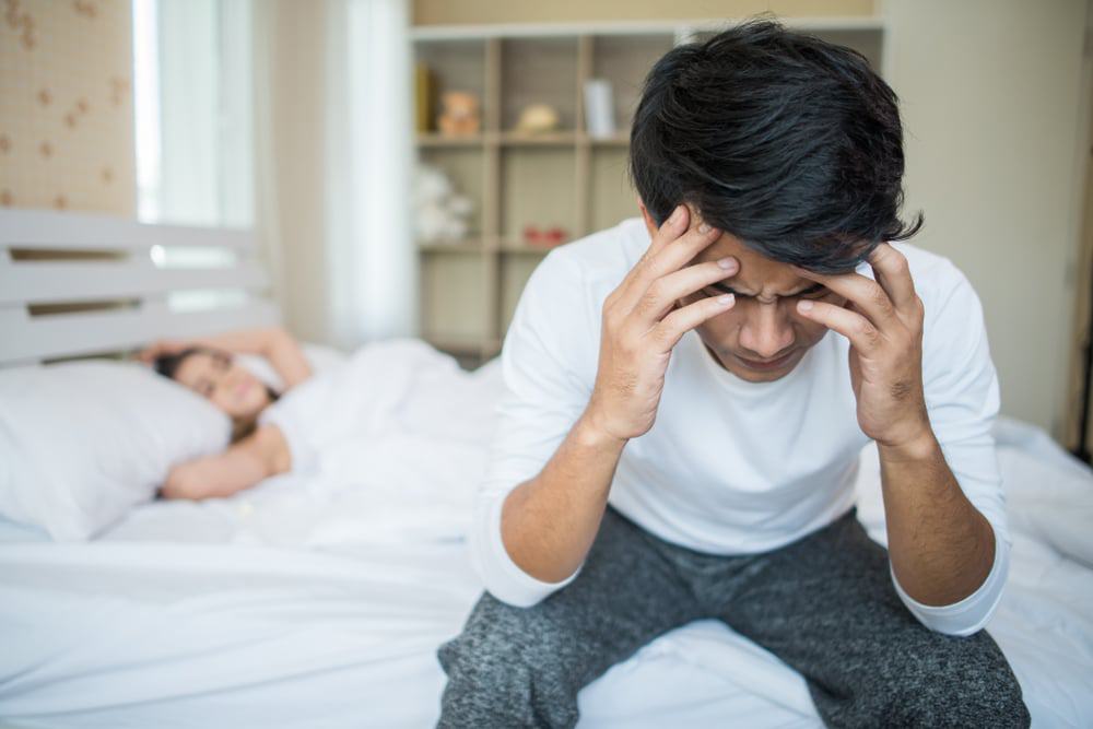 Homem sentado na cama com as mãos na cabeça enquanto mulher está deitada ao fundo