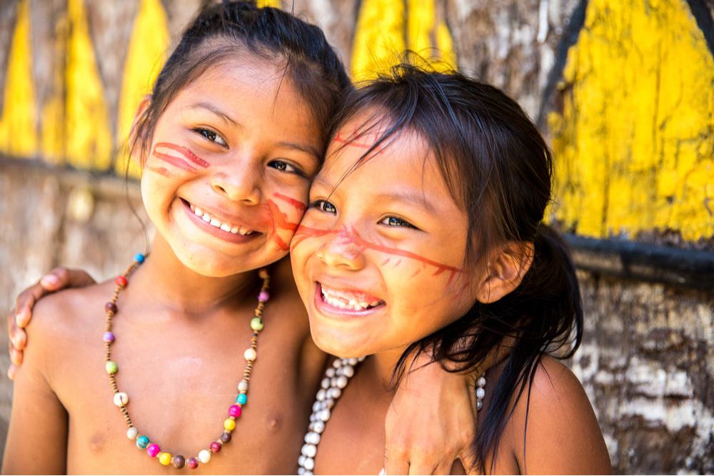 Imagem mostra duas crianças indígenas sorridentes e abraçadas, com pinturas faciais e vestimentas nativas em evidência
