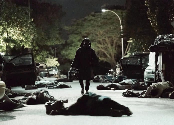 Foto mostra personagem utilizando capa e máscara de proteção biológica enquanto caminha por rua escura repleta de corpos pelo chão.