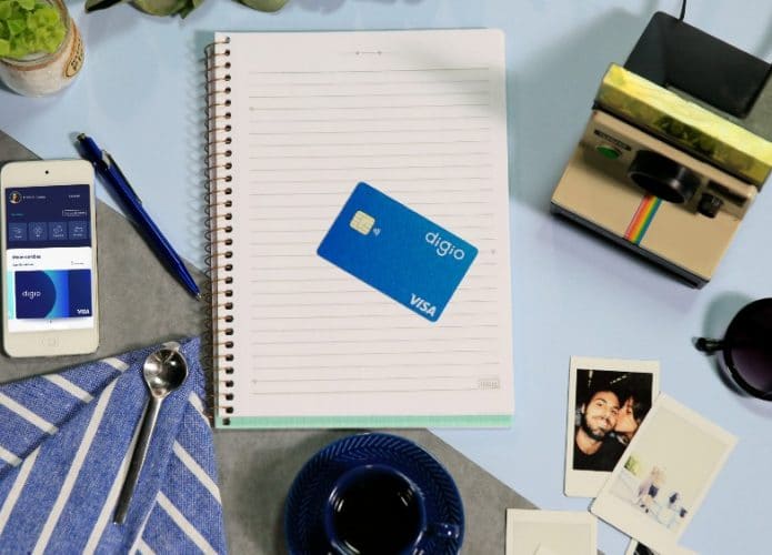 Imagem mostra um cartão do banco digital Digio em cima de um caderno pautado; ao redor há elementos de uma mesa de escritório e a foto foi tirada de uma perspectiva vista de cima