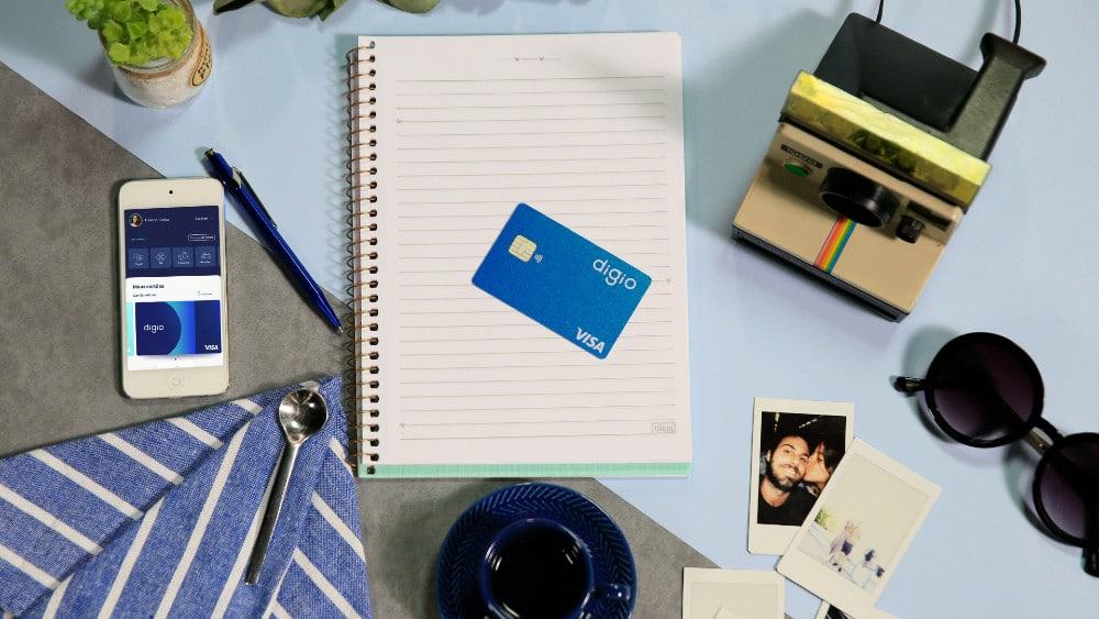 Imagem mostra um cartão do banco digital Digio em cima de um caderno pautado; ao redor há elementos de uma mesa de escritório e a foto foi tirada de uma perspectiva vista de cima
