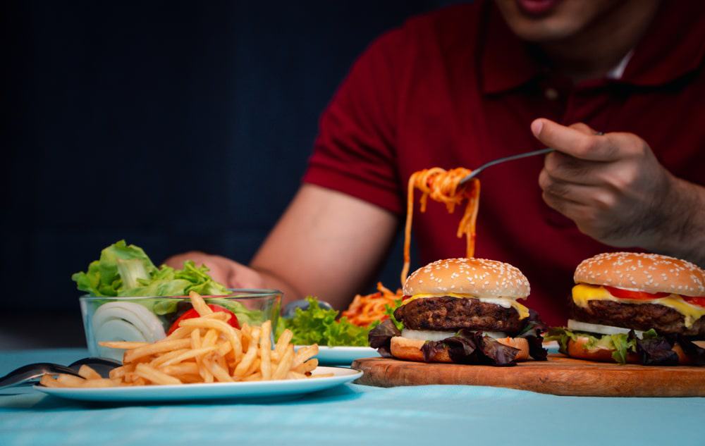 Homem sentando em frente a uma mesa com batatas fritas, dois hambúrgueres, salada e macarronada  