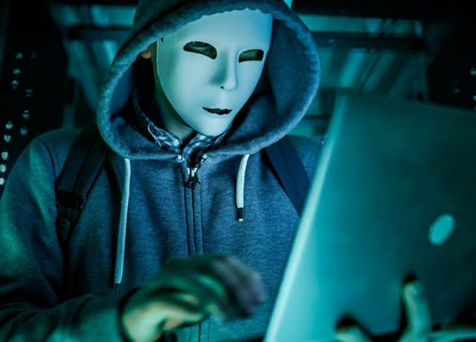 hacker com máscara e moletom de capuz digitando em notebook