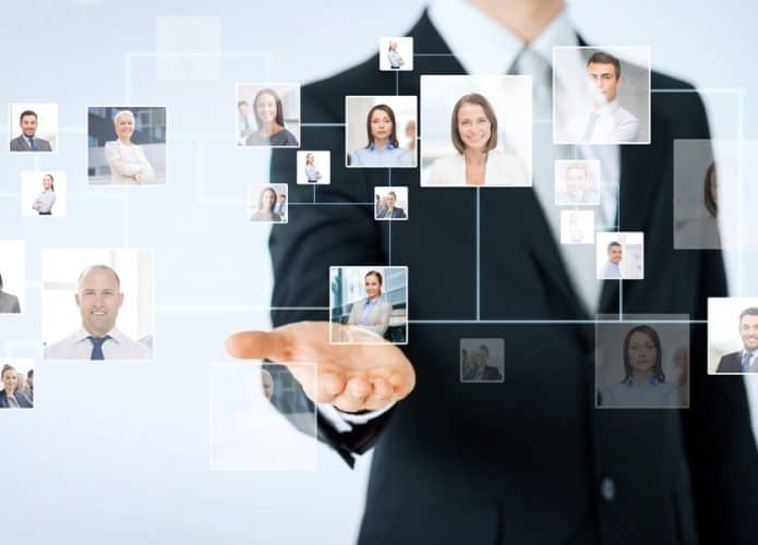 Imagem mostra o torço de um homem com traje formal e a mão direita levantada com a palma para cima; à frente, pode-se ver diversas fotos 3x4 digitalizadas, de pessoas, como se o homem estivesse usando uma tecnologia para selecionar uma dessas pessoas