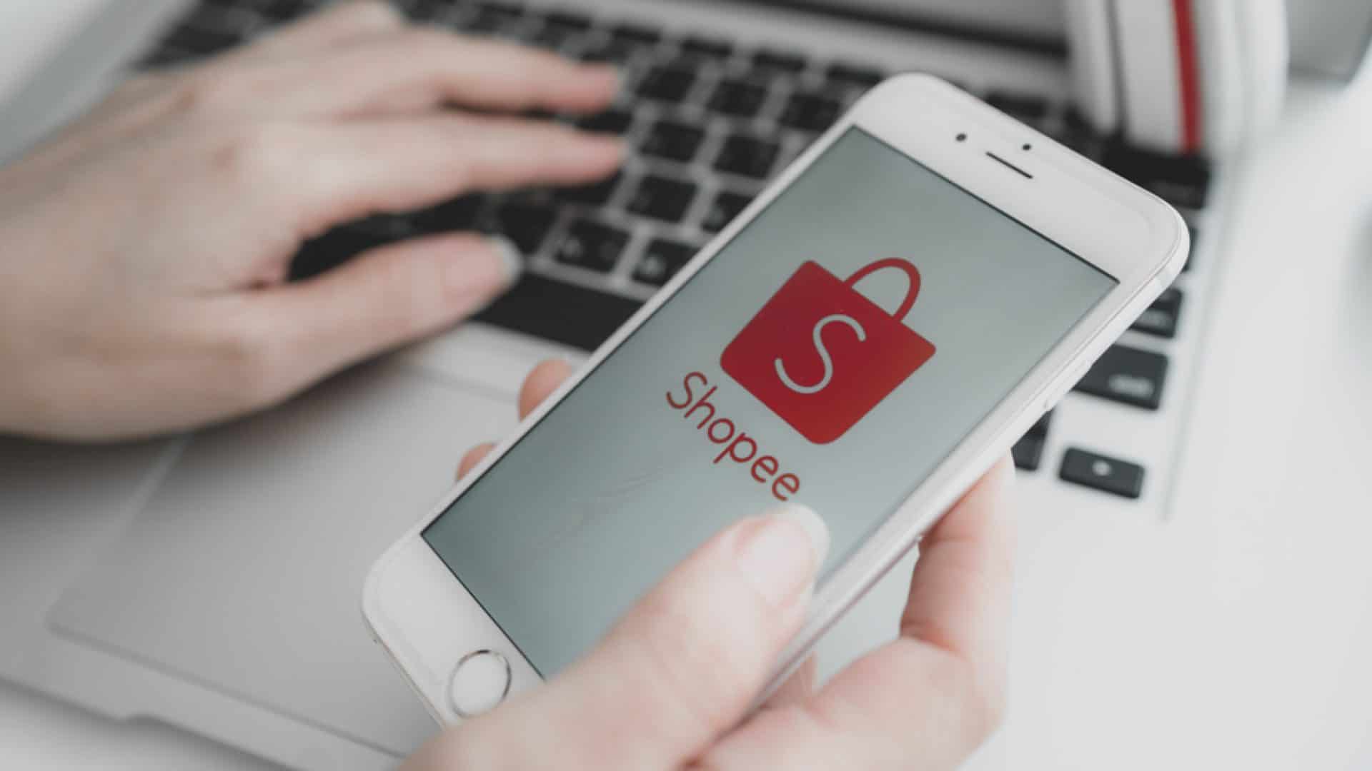Imagem mostra uma mão segurando um smartphone, na tela aparece o logotipo do e-commerce Shopee. Ao fundo há uma mãe digitando algo em um teclado de notebook.