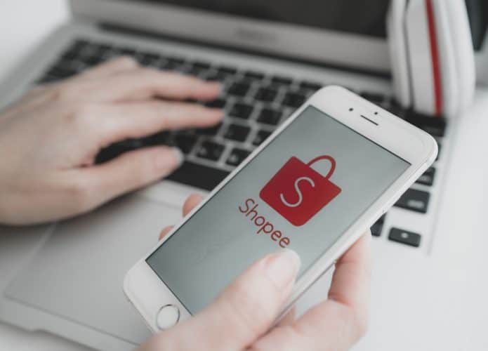 Imagem mostra uma mão segurando um smartphone, na tela aparece o logotipo do e-commerce Shopee. Ao fundo há uma mãe digitando algo em um teclado de notebook.