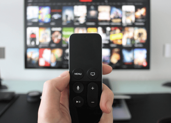 Imagem de uma mão segurando um controle remoto, com uma TV ao fundo exibindo vários programas