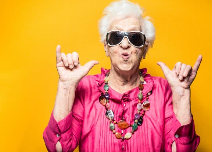 Senhora de idade vestindo trajes coloridos e óculos escuros, fazendo uma pose mais jovial à frente de um fundo alaranjado. A ideia da imagem é passar longevidade e juventude mesmo em idades avançadas
