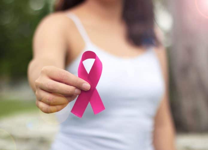 Imagem mostra o torso de uma mulher ao fundo, à frente ela segura um laço rosa, símbolo da campanha Outubro Rosa, de prevenção ao câncer de mama