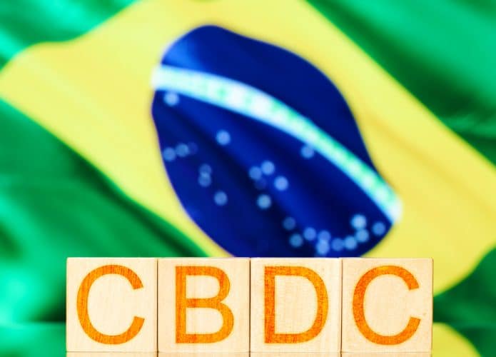 Cubos de madeira com a inscrição CBDC (Real Digital) com a bandeira do Brasil ao fundo