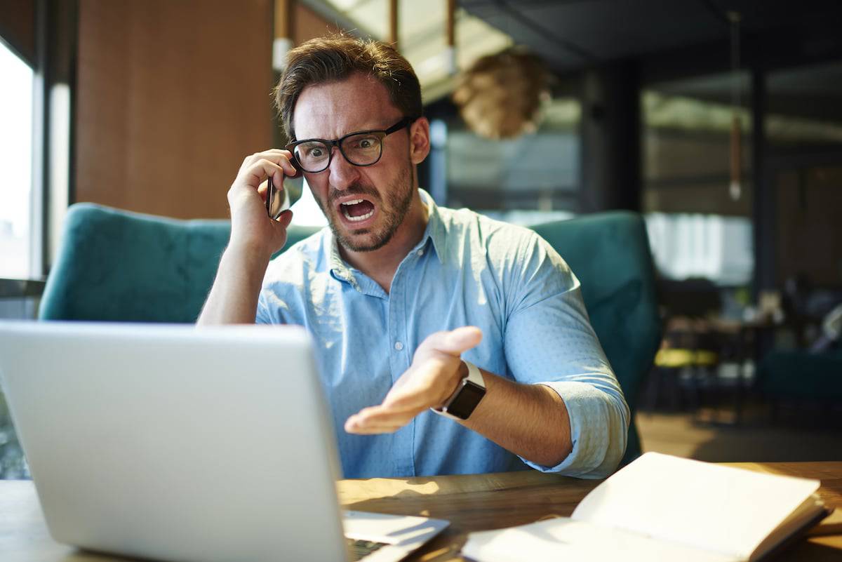 imagem mostra homem irritado e gritando, sentado e de frente para um notebook, enquanto fala ao celular