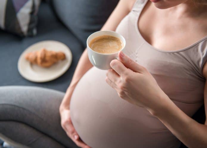 Mulher grávida tomando café, sentada no sofá e passando a mão na barriga