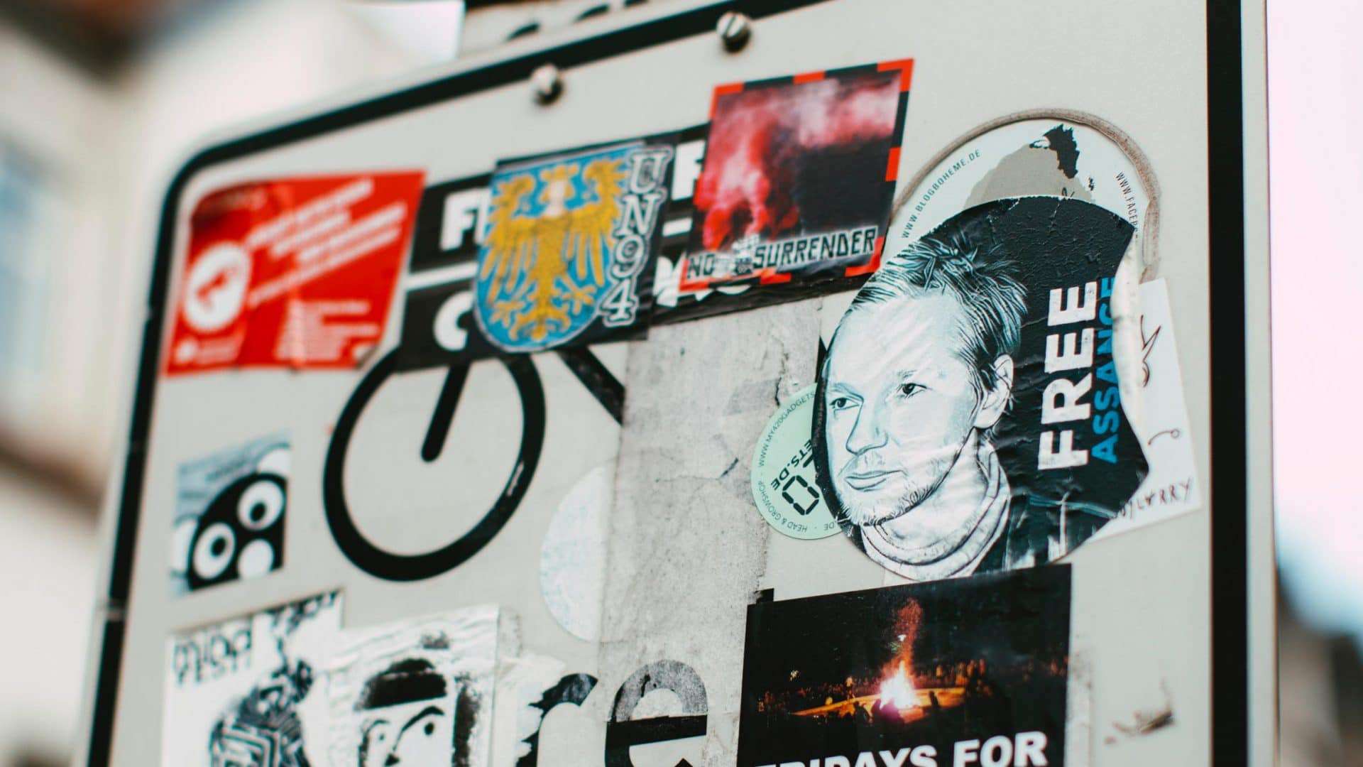 placa de trânsito coberta com adesivos em que um deles é pedindo a favor de Julian Assange
