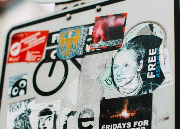 placa de trânsito coberta com adesivos em que um deles é pedindo a favor de Julian Assange