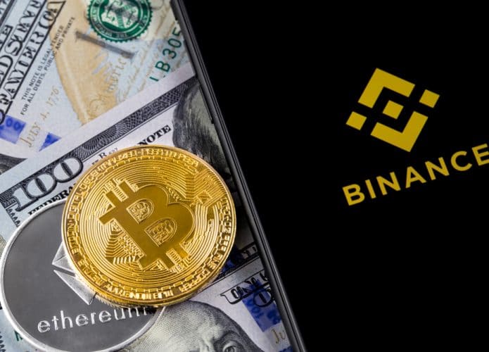 iPhone exibindo o logotipo da Binance na tela com uma moeda de bitcoin, outra de ethereum e notas de dólares ao fundo. Binance é uma corretora de criptomoedas