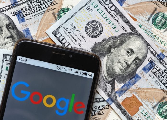 Logotipo do Google é exibido na tela de um smartphone com notas de 100 dólares ao fundo