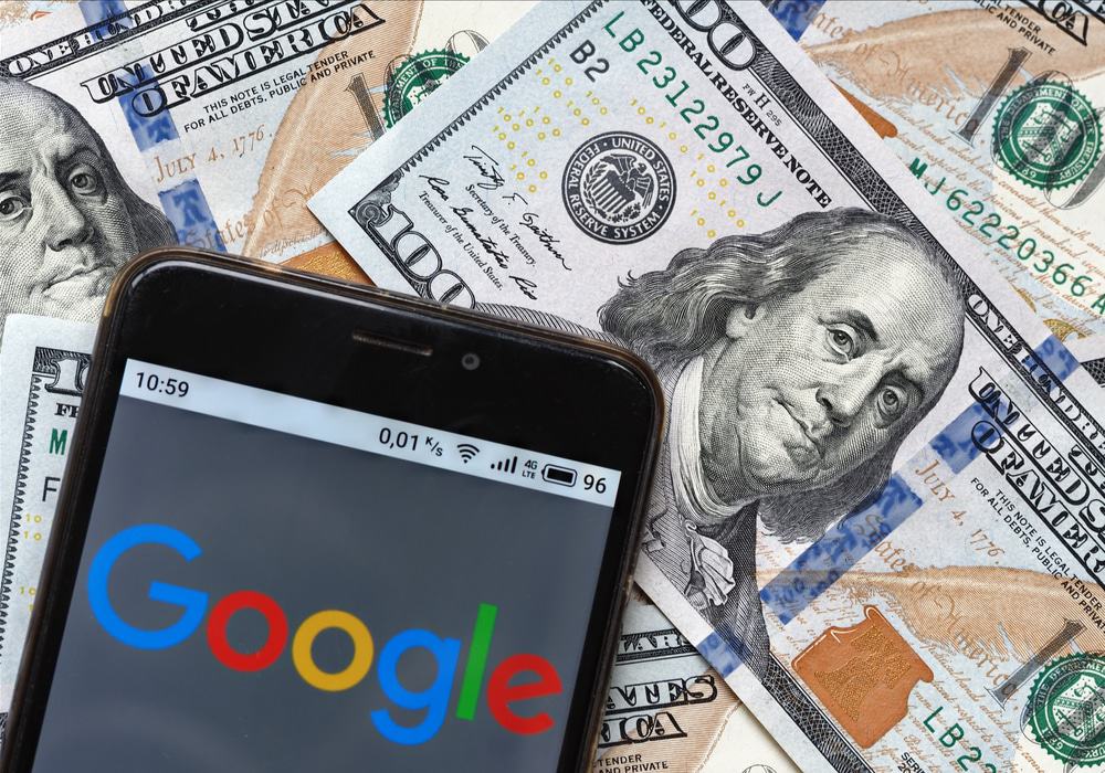 Logotipo do Google é exibido na tela de um smartphone com notas de 100 dólares ao fundo