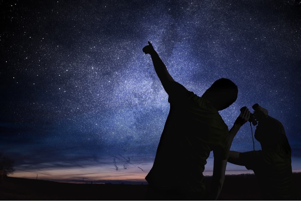 Imagem mostra duas silhuetas humanas observando um céu estrelado com um binóculo