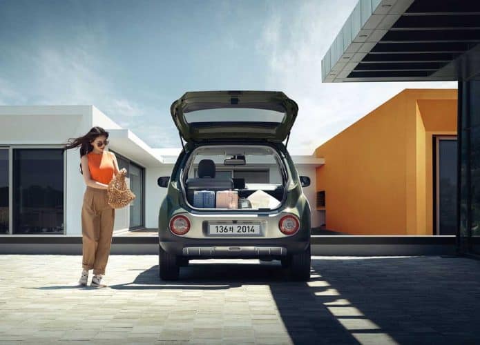 Uma mulher segurando uma bolsa e andando ao lado de uma pequena van Hyundai Casper com o porta-malas aberto e cheio de compras