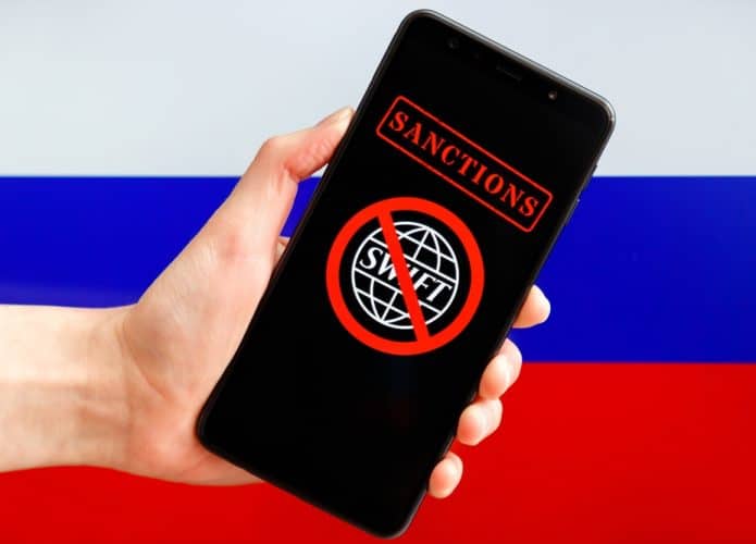 Mão segurando celular exibindo logotipo do sistema financeiro SWIFT sob sinal vermelho de proibição com bandeira russa ao fundo. Sanções contra a Rússia, desconexão do SWIFT