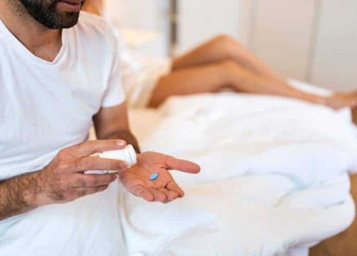 Homem sentado na cama colocando um comprimido azul na mão enquanto uma mulher está deitada ao fundo
