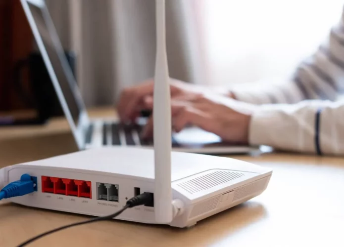 Pessoa teclando em notebook ao lado de um roteador numa mesa