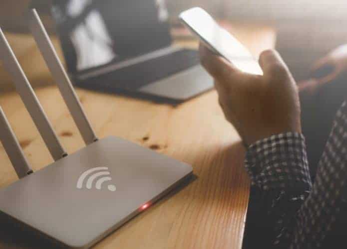 Pessoa segurando um smartphone ao lado de um roteador e notebook na mesa