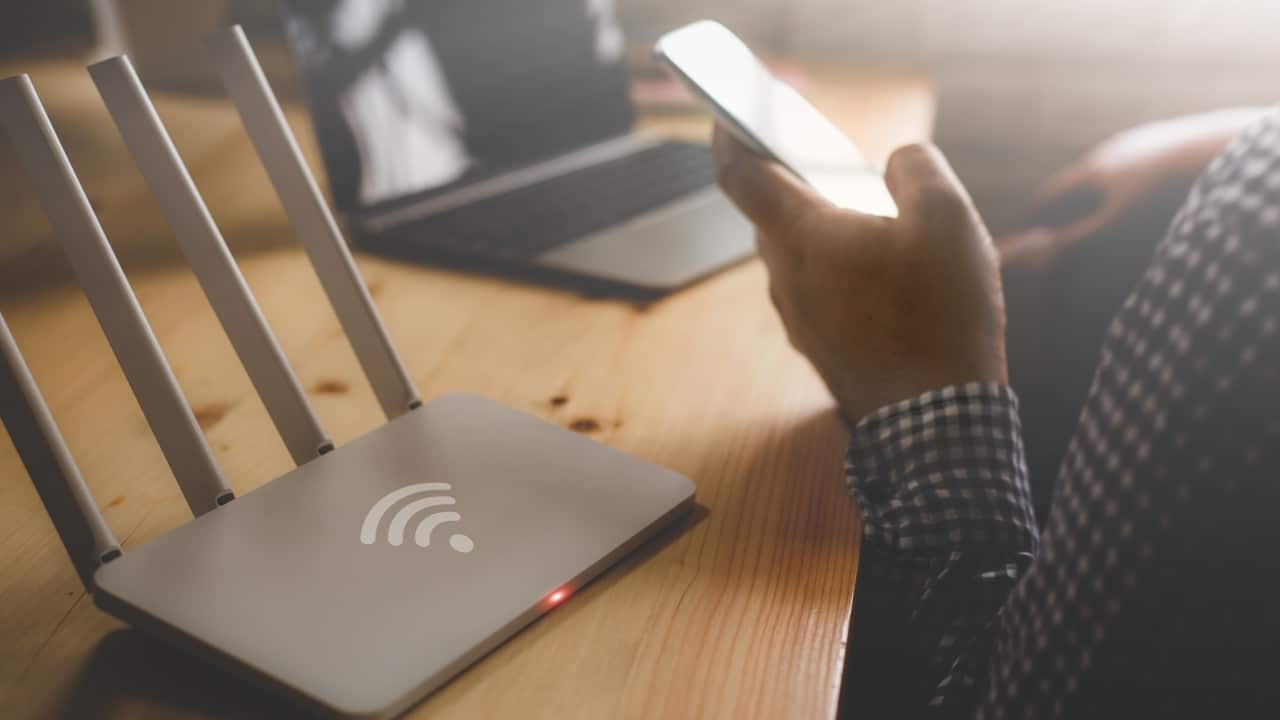 Pessoa segurando um smartphone ao lado de um roteador e notebook na mesa