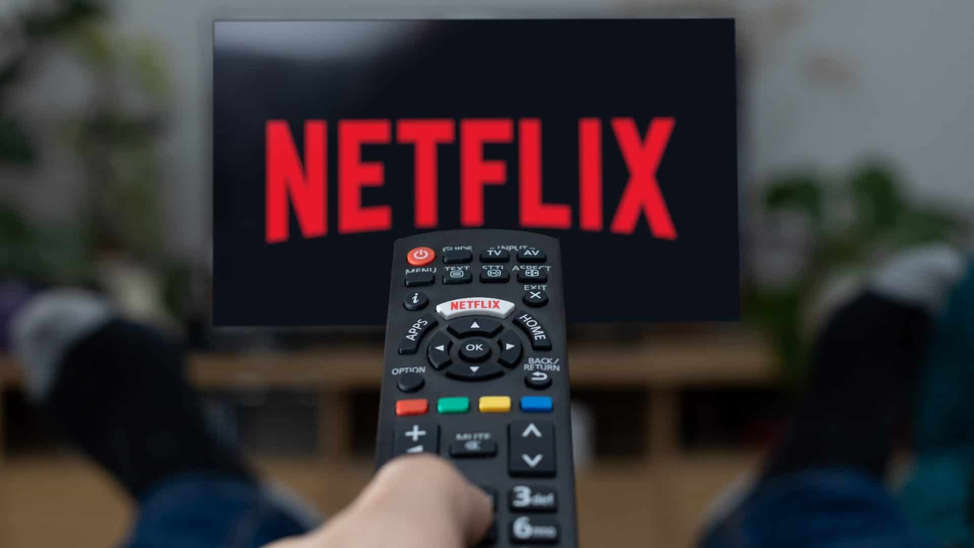 Pessoa apontando controle para TV, que exibe logomarca da Netflix na tela