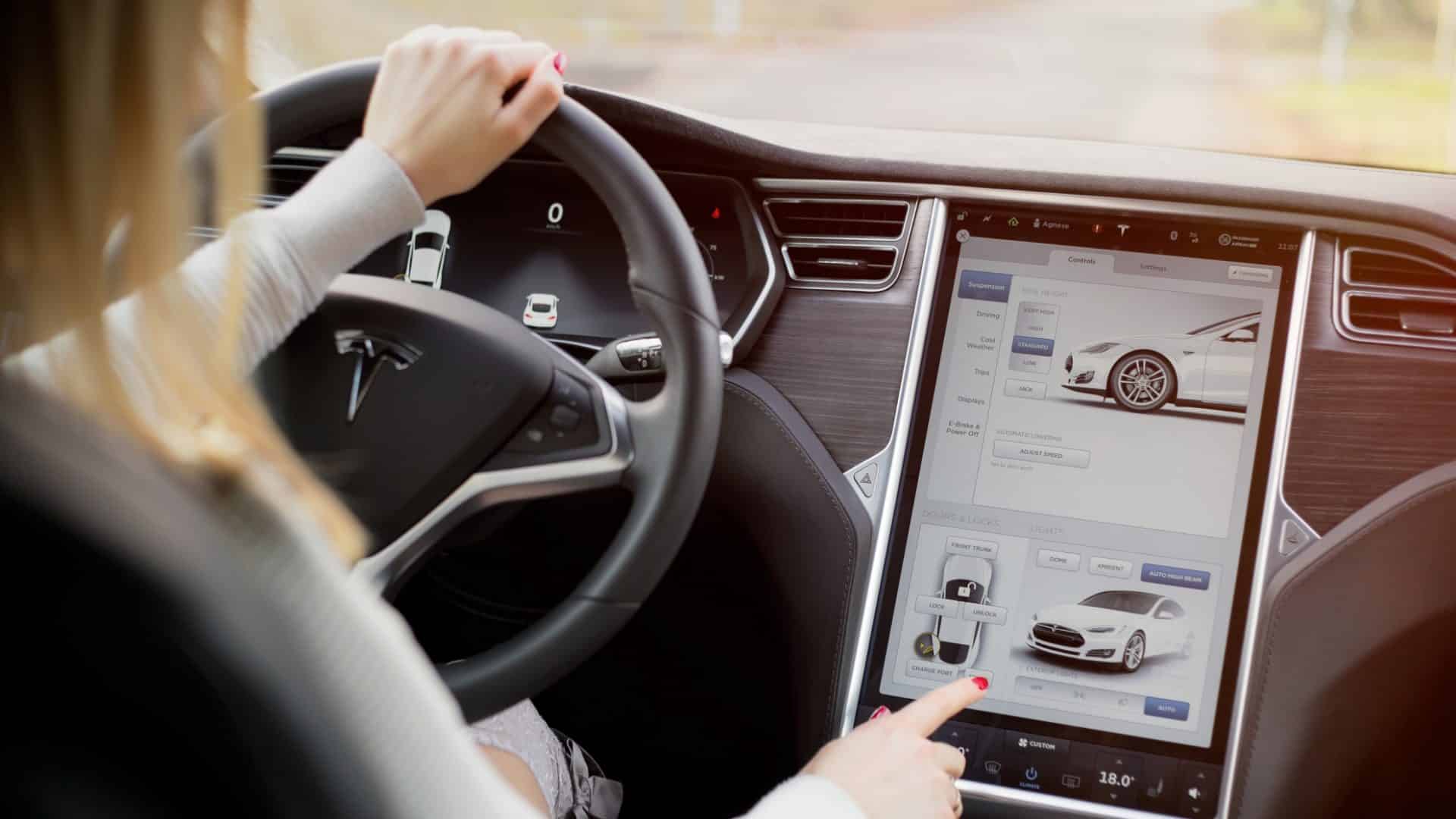 Interior de um veículo elétrico Tesla Model S; mulher dirigindo e tocando no painel touchscreen.